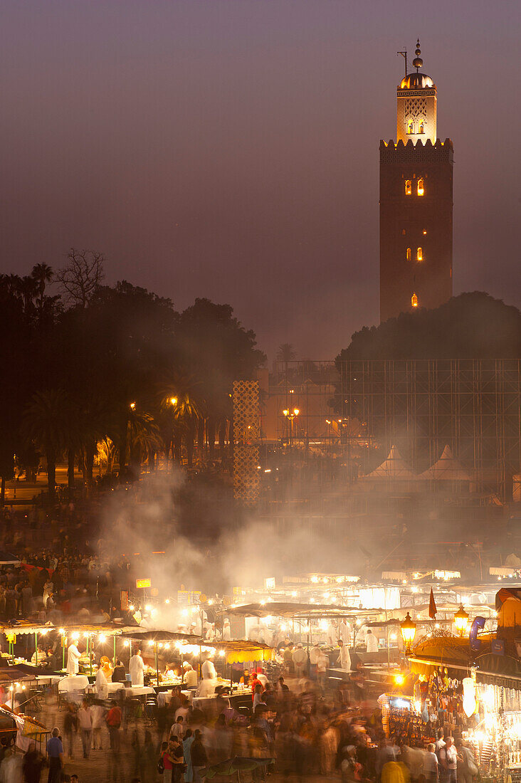 Looking across food stalls in Dejmaa el Fna at dusk with minaret of Koutoubia Mosque behind, Marrakesh, Morocco