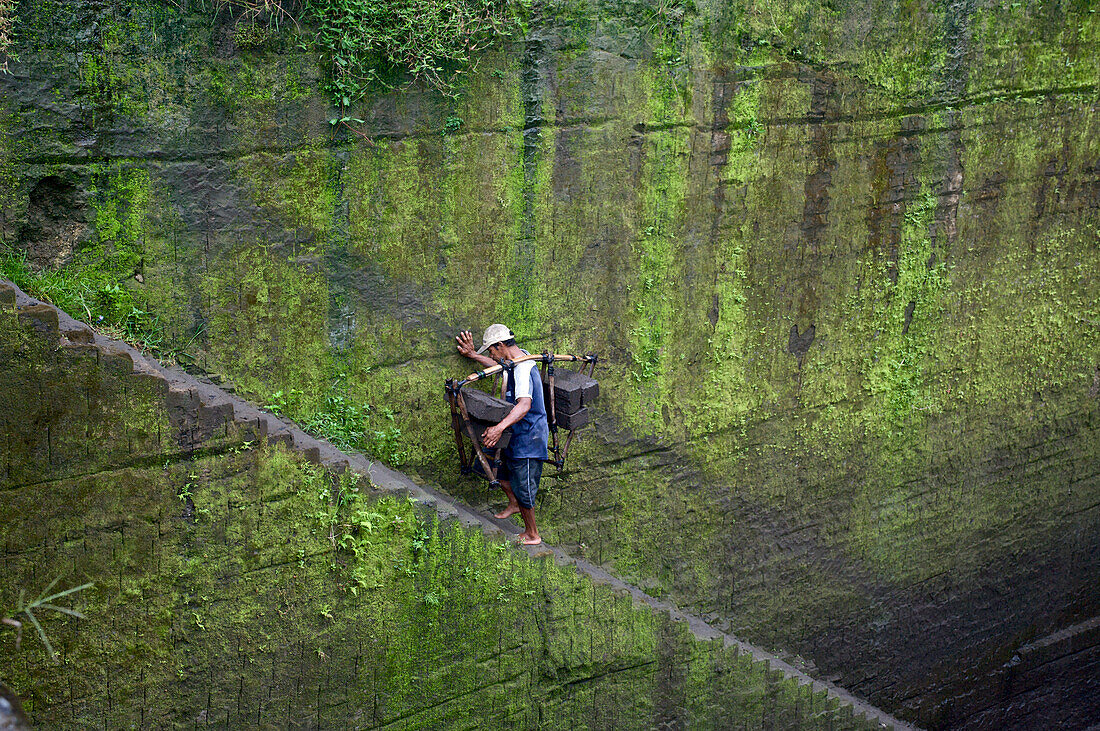 Javanischer Arbeiter trägt Steine auf einer ungesicherten Treppe aus einer Grube in einem Steinbruch, Bali, Südwestküste, Indonesien