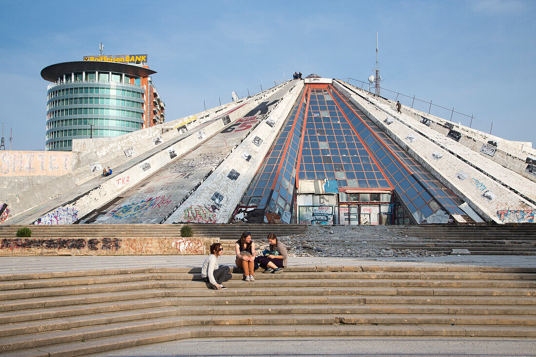 People sitting on the steps of the graffiti-covered The Pyramid International Center of Culture with glass high-rise building of Austrian Raiffeisen Bank behind, Tirana, Albania