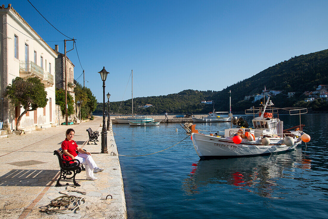 Woman sitting on a bench in the idyllic fishing village with fishing boat, Kioni, Ithaca, Ionian Islands, Greece