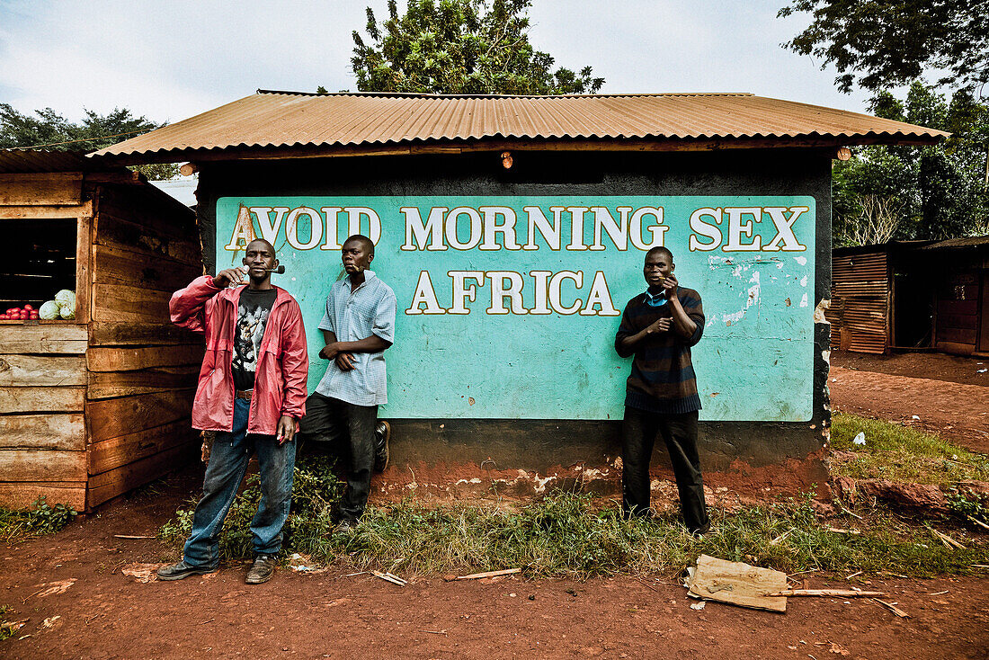 Three men smoking pipes in front of a hut with Anti-Aids campaign, Buwenda, Uganda, Africa