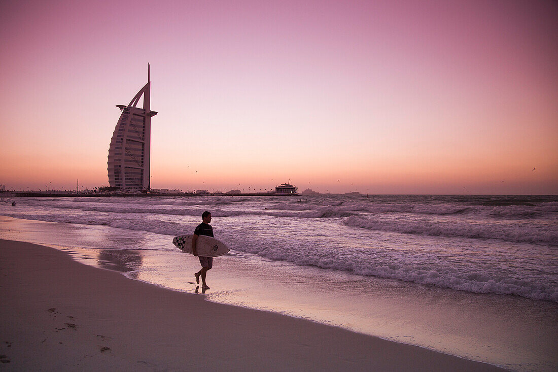 Surfer läuft entlang Strand nahe Burj al Arab Hotel bei Sonnenuntergang, Dubai, Vereinigte Arabische Emirate
