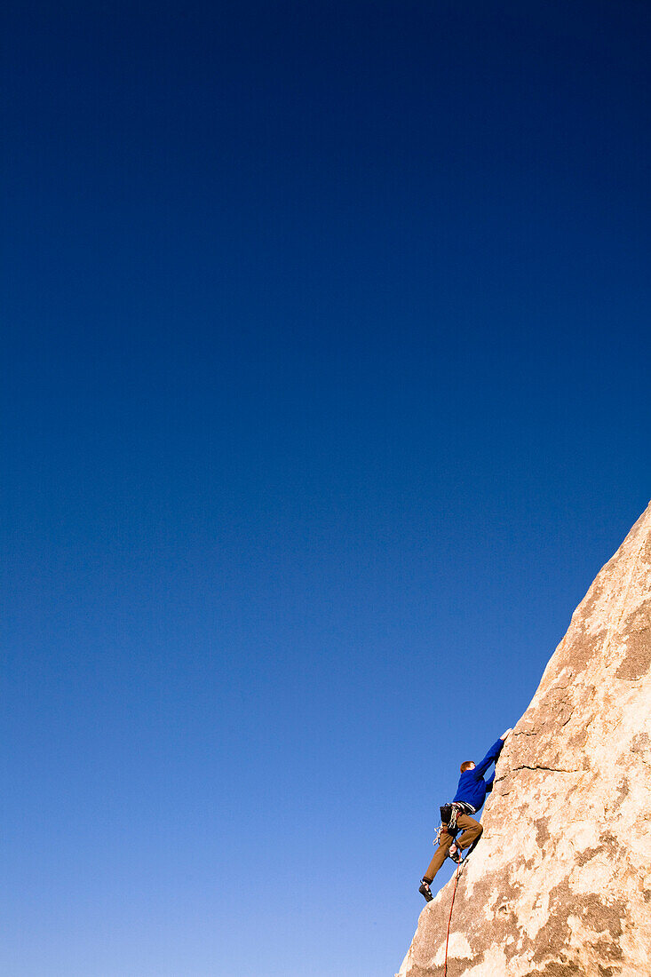 A climber in a blue jacket reaches for a handhold while climbing the Southwest Corner of Headstone Rock in Joshua Tree National Park, California Joshua Tree, California, United States of America