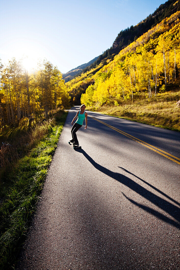 A young woman longboards down a smooth country road through the mountain peaks and gold forests Aspen, Colorado, USA