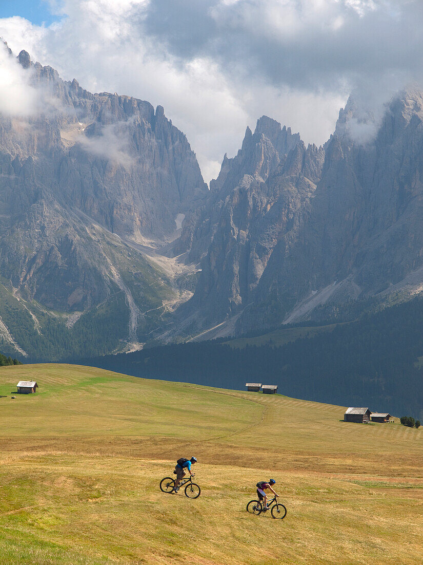 Two mountain bikers are riding downhill a grassy slope at Seiser Alm, with rock cliffs in the background Val Gardena, Dolomites, Italy