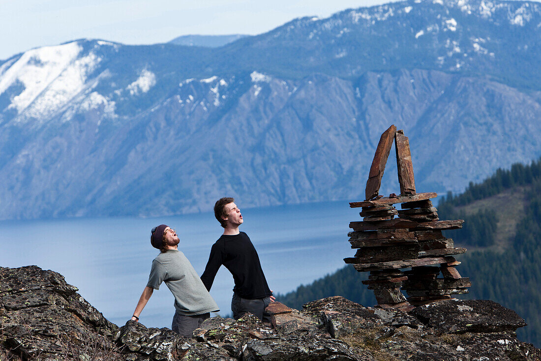 Two young men howl next to a balanced rock stack above a mountain lake in Idaho Sandpoint, Idaho, USA