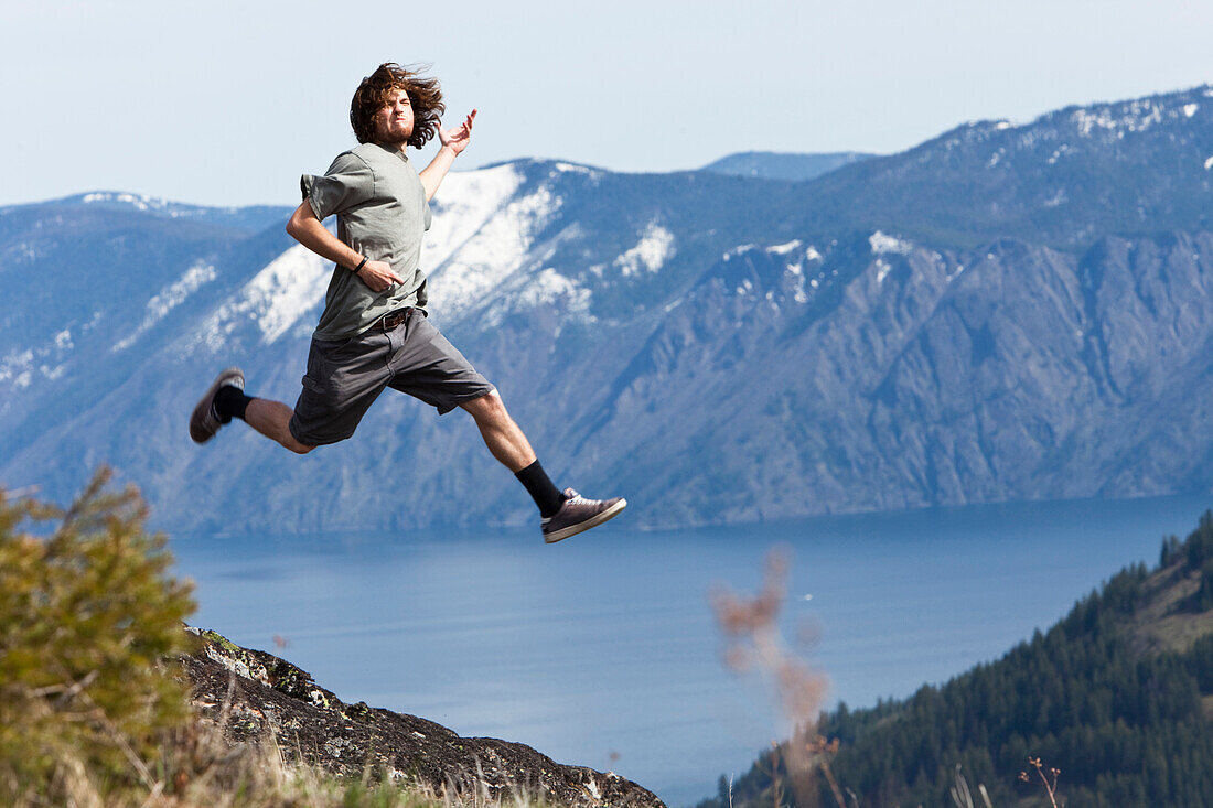 A young man grins as he jumps and does a air guitar above a mountain lake in Idaho Sandpoint, Idaho, USA