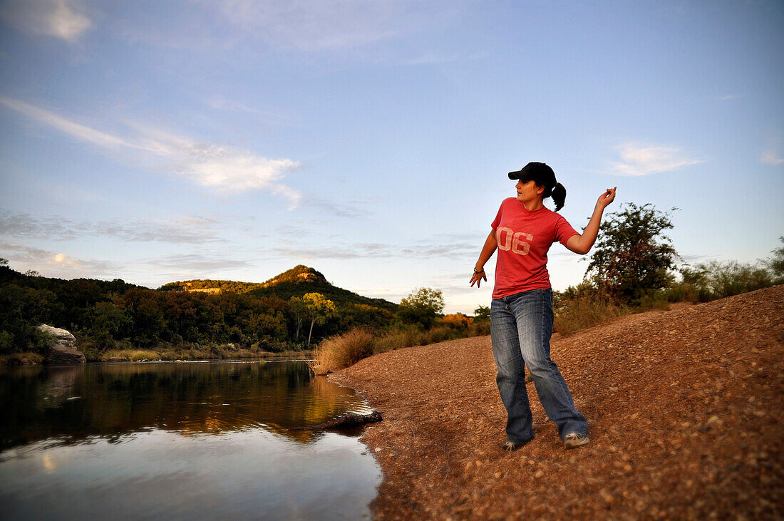 A young woman playfully throws rocks into a river at sundown