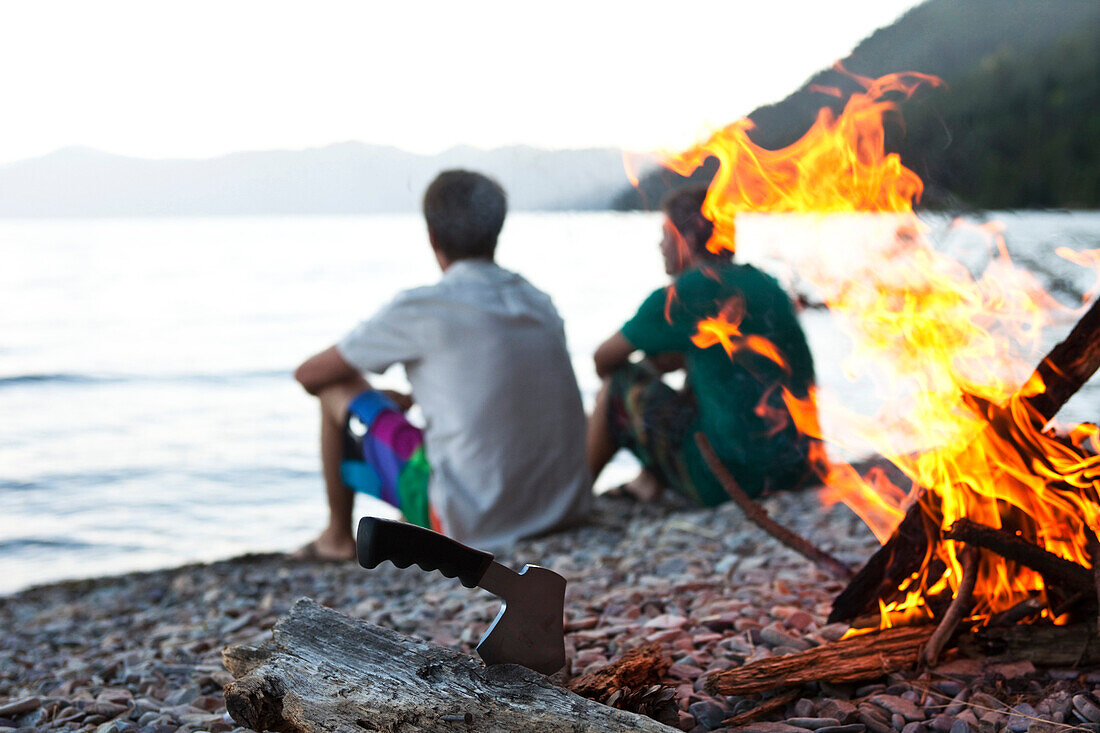 Two young men sit next to a campfire next to a lake on a camping trip in Idaho., Sandpoint, Idaho, USA