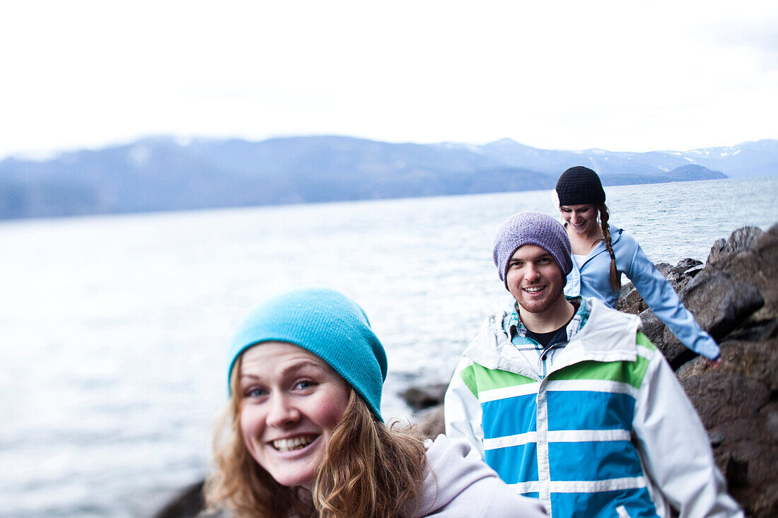 A group of three young adults smile while hiking at the edge of a lake in Idaho., Sandpoint, Idaho, USA