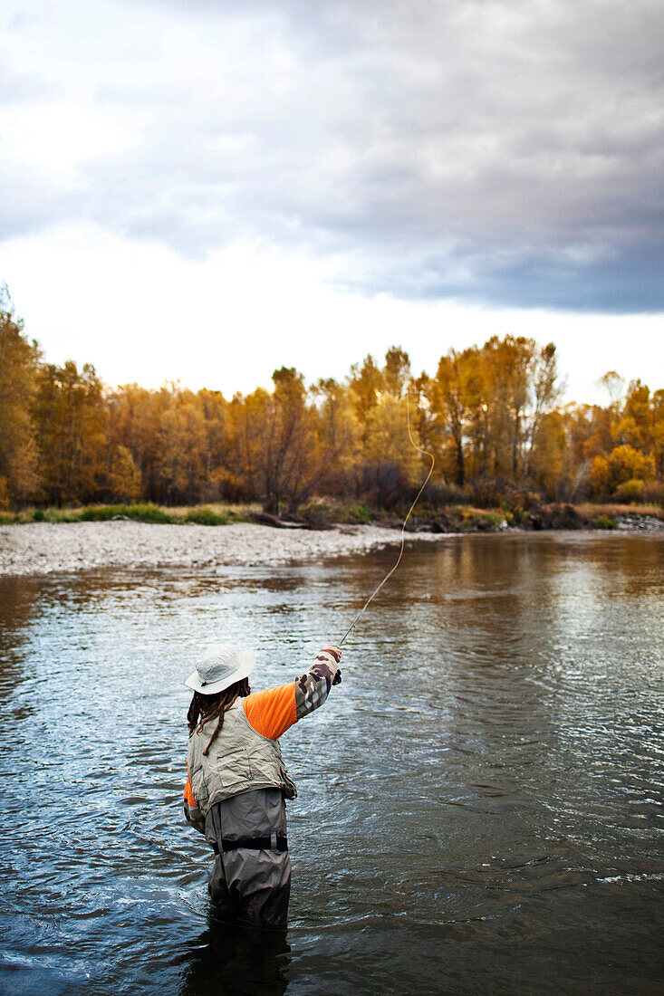 A athletic man fly fishing stands in a river with the fall colors and snowy mountains behind him., Bozeman, Montana, USA