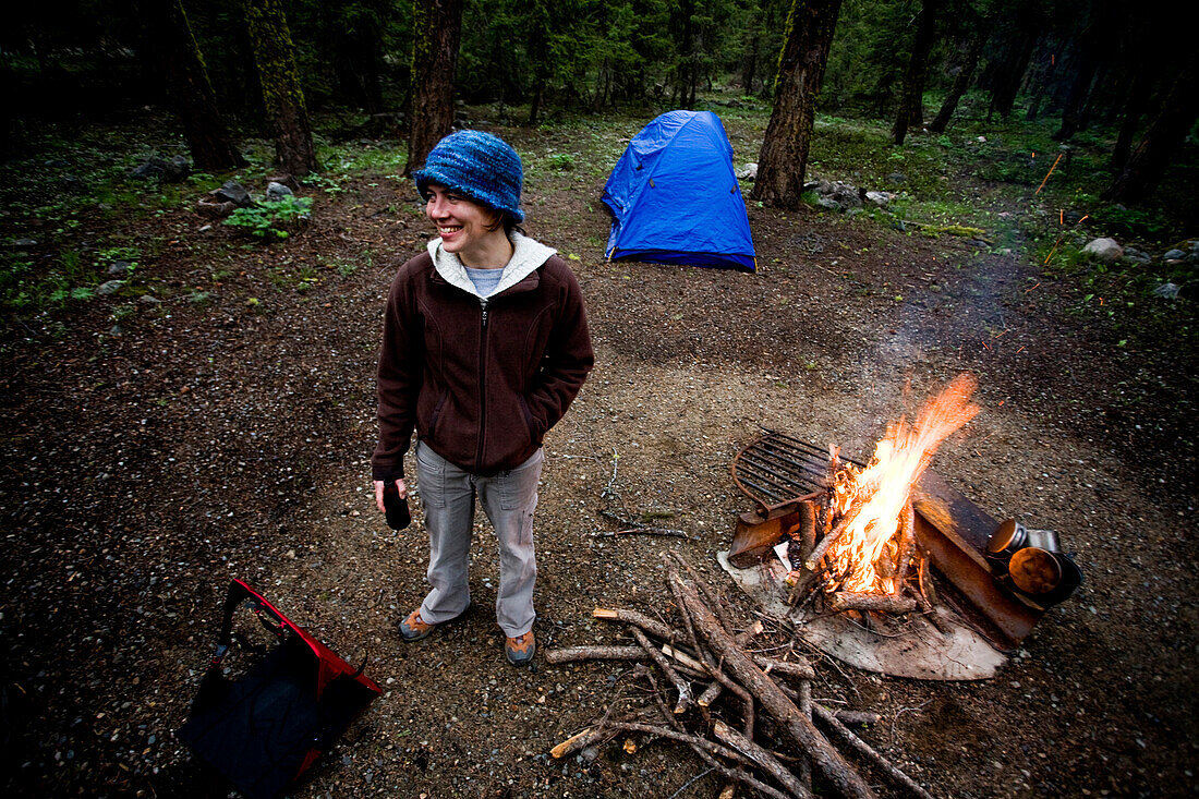 A young woman stands near a campfire with a tent in the background Mazama, Washington, USA