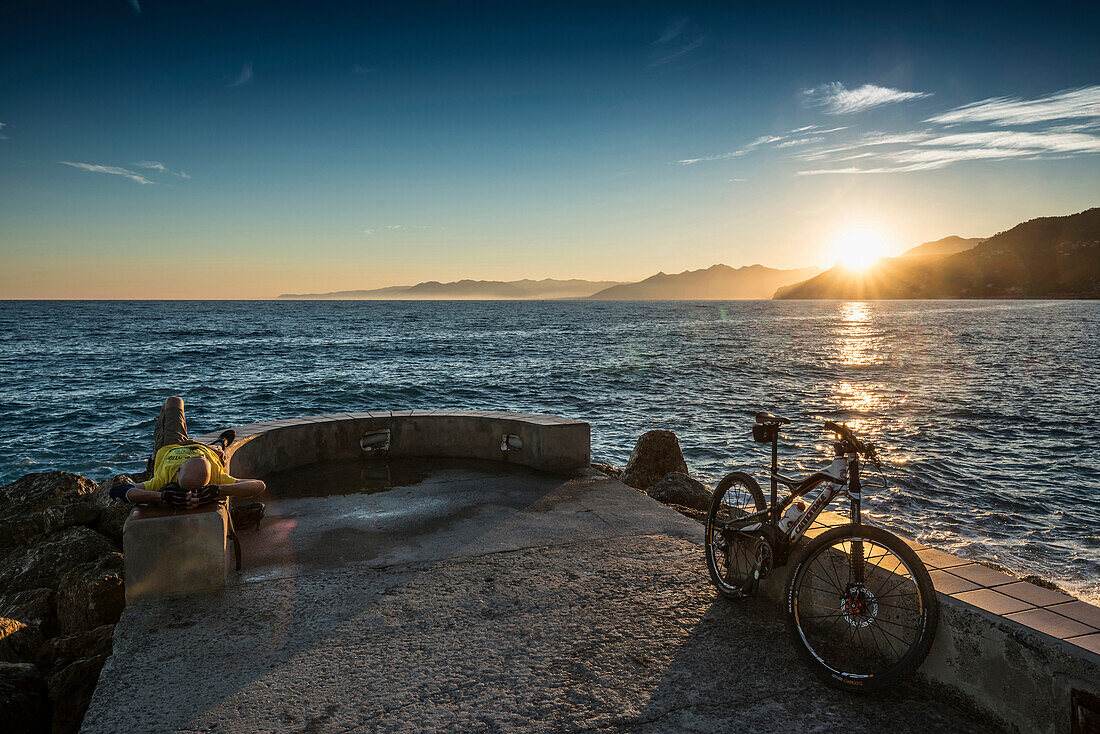 Mountain biker resting on quay wall in sunset, Varigotti, Finale Ligure, Province of Savona, Liguria, Italy