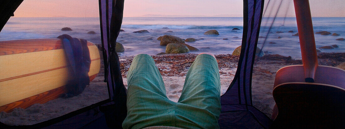 A young man lying in his tent with a guitar while camping on the beach waiting for surf Carpinteria, California, USA