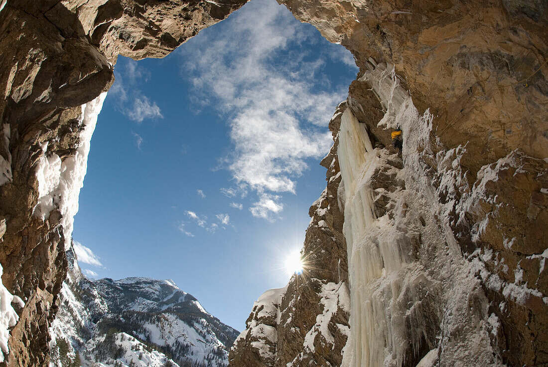 View from below as a professional male ice climber ascends a frozen waterfall on a sheer rock face Ouray, Colorado, USA