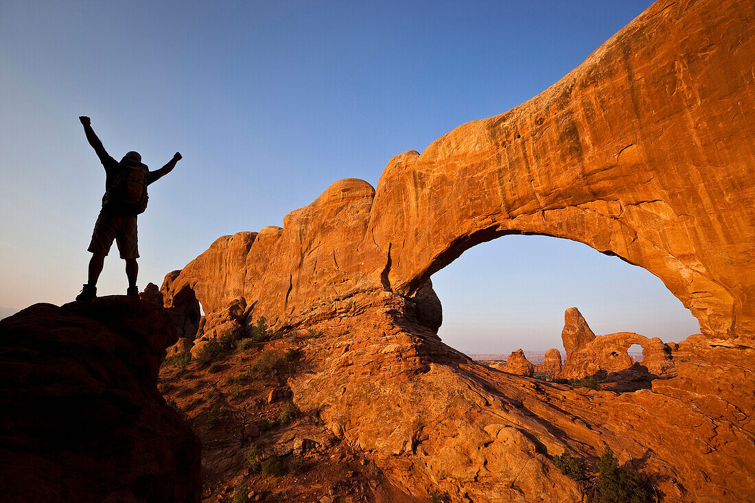 A silhouetted hiker raises his arms beside an arch in Arches National Park, Utah Moab, Utah, USA