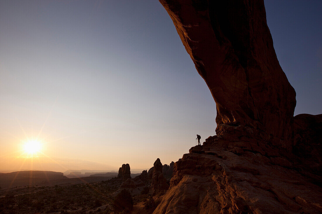 A hiker stands beneath an arch looking at the sunrise in Arches National Park, Utah Moab, Utah, USA