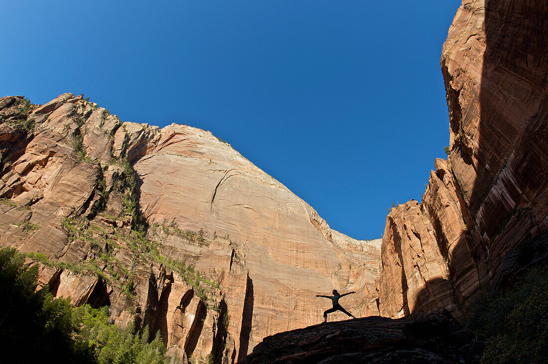 A young woman practicing yoga on a rock is silhouetted against distant cliffs Zion, Utah, USA