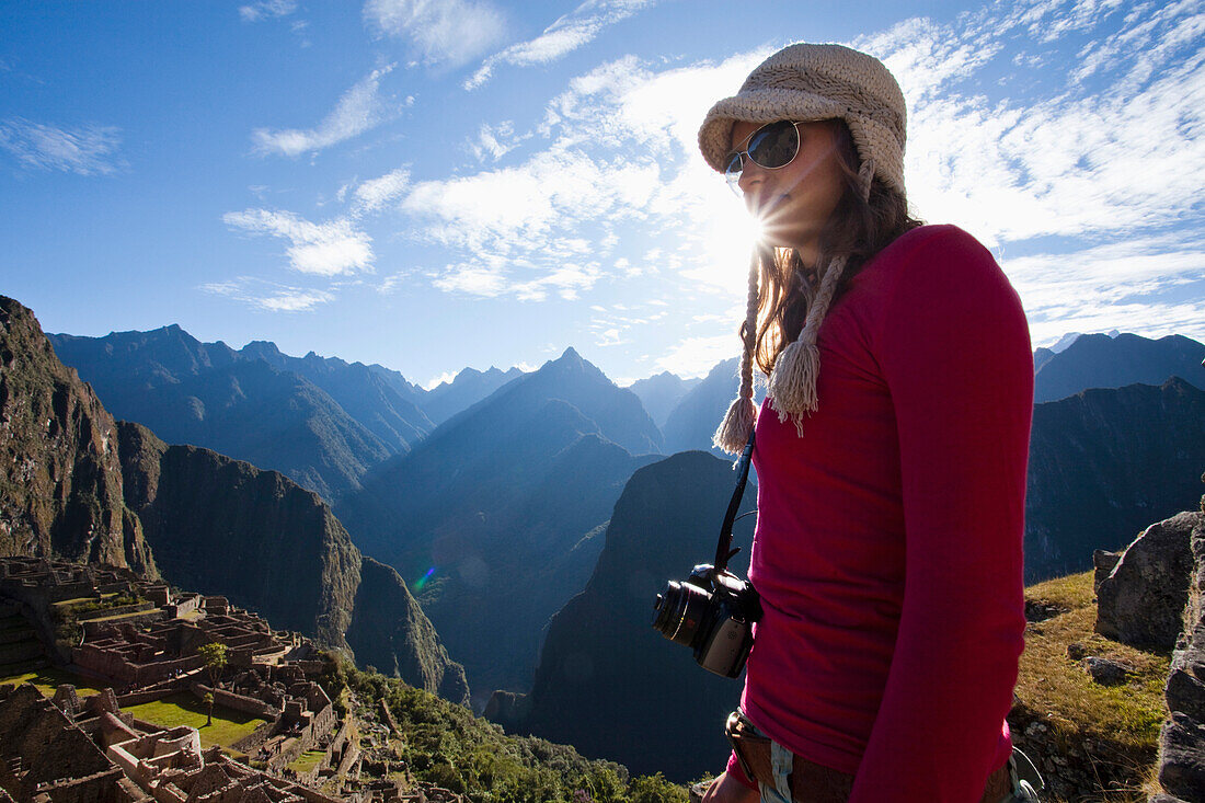 A young girl stands with a camera around her neck overlooking a steep valley, the rising sun, and the ruins of a lost civilization Machu Picchu, Peru