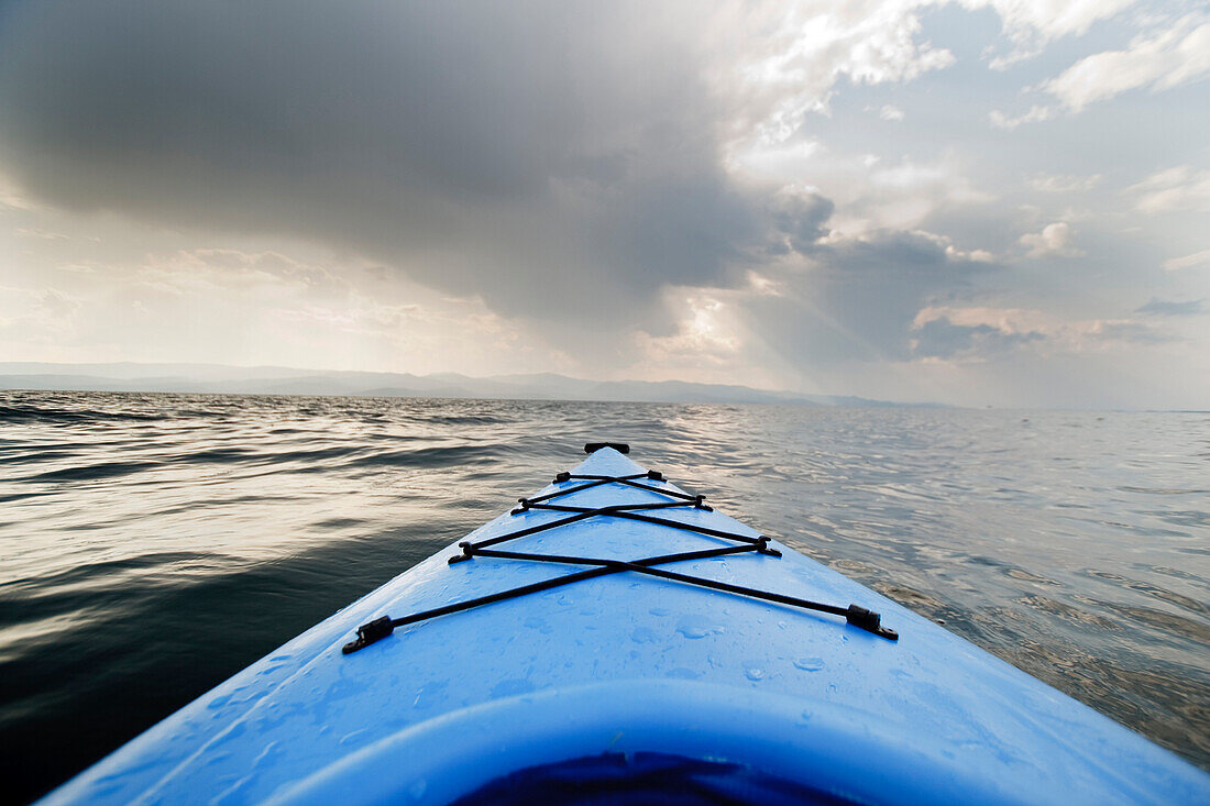 The front of a blue kayak floats across the water of Flathead Lake, Montana, as an evening storm builds in the distance Bigfork, Montana, USA