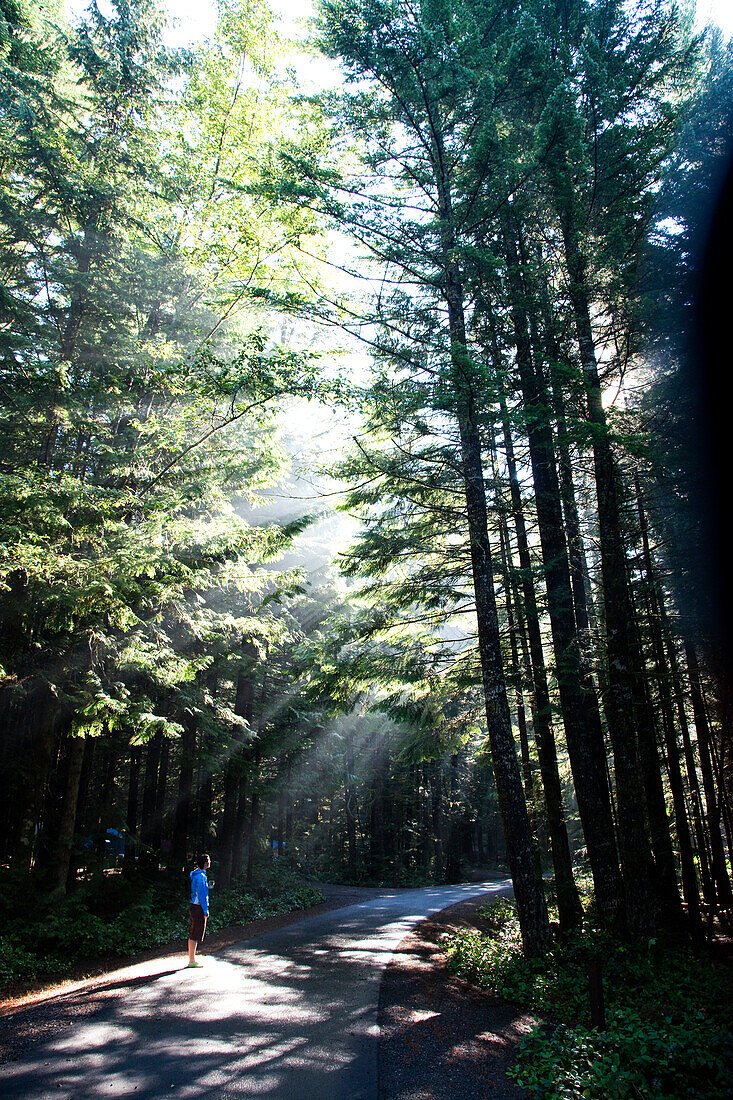 A young woman wearing a blue top walks down an empty road as the suns filters through the trees and mist Squamish, British Columbia, Canada