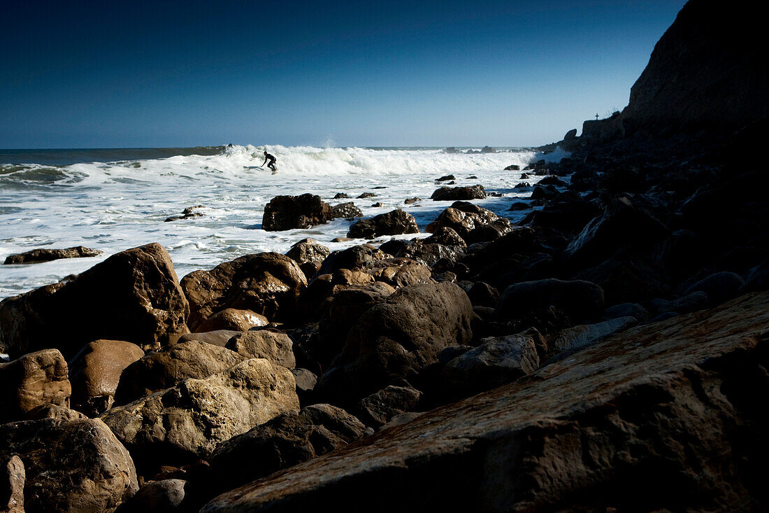 One male surfer riding a wave over a shallow reef with cliffs and a cross in the background Ventura, California, USA