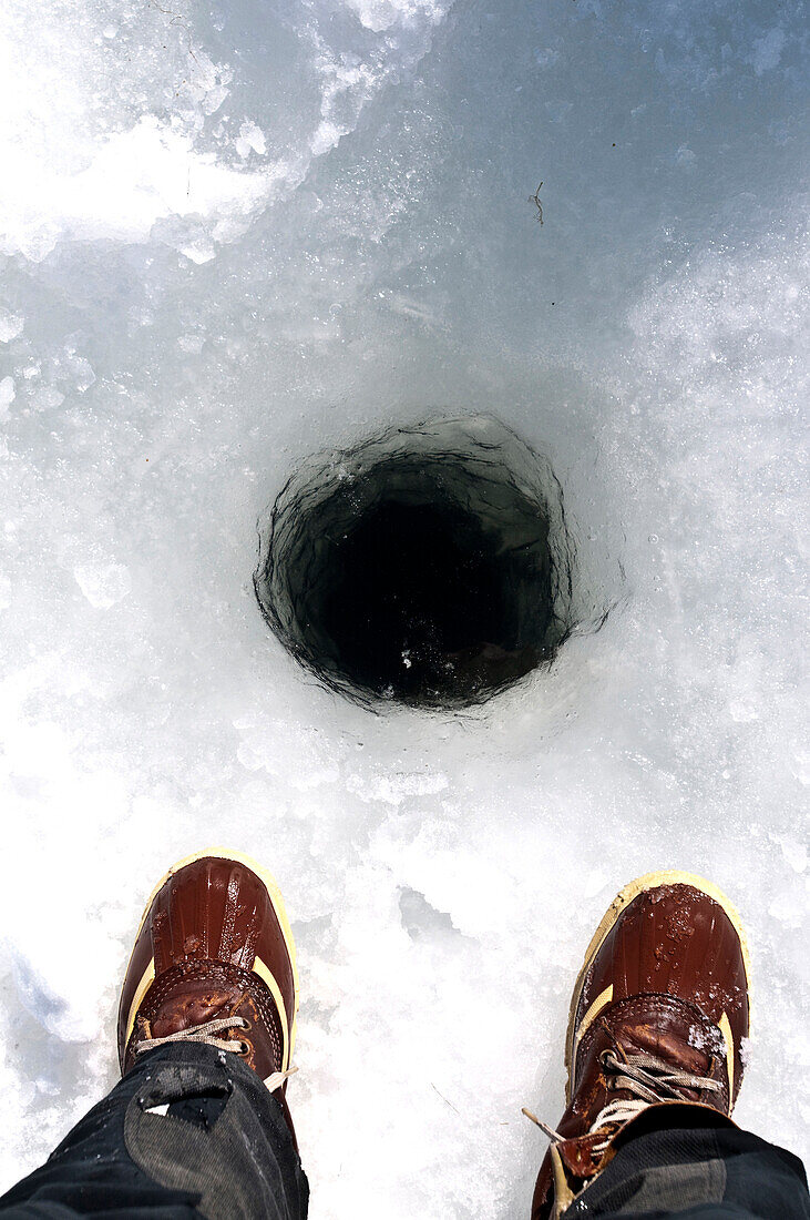 Ice fisherman's boots looking down into a dark hole in the ice Tamworth, New Hampshire, USA