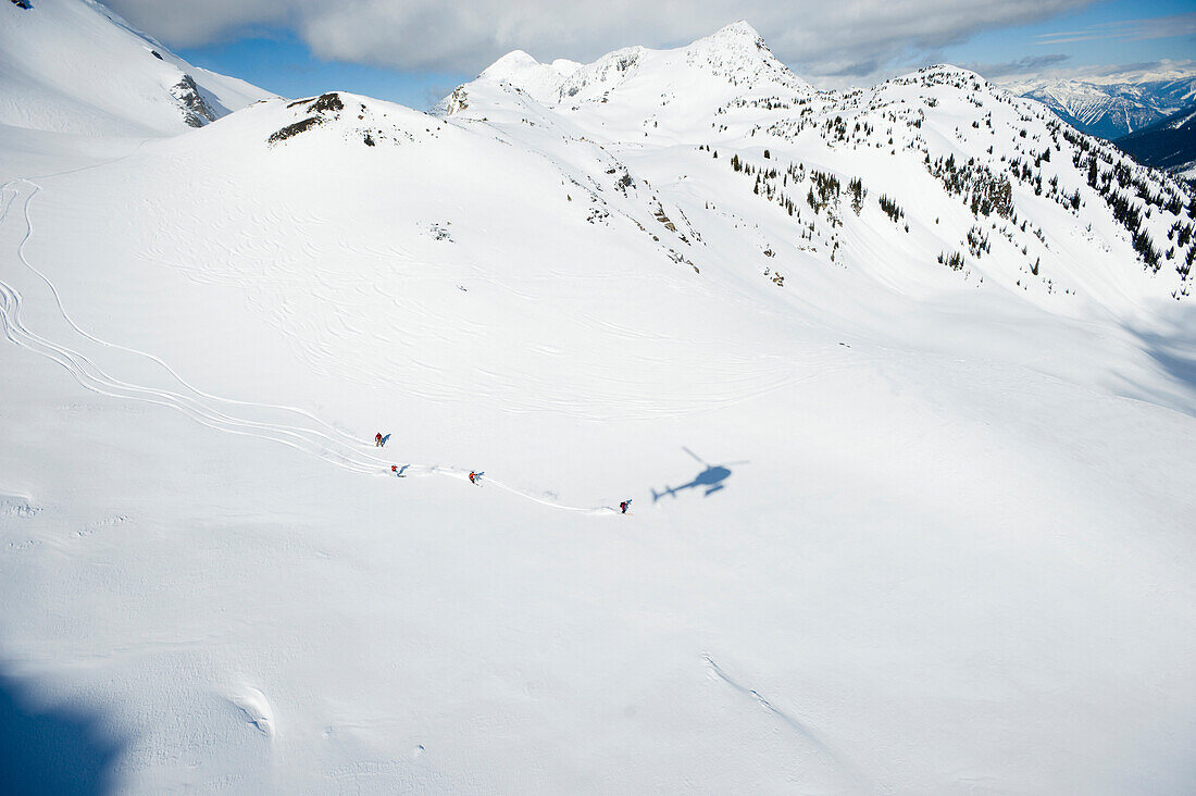 A group of skiers take a run down a large alpine bowl in the backcountry while the shadow of their helicopter is follows British Columbia, Canada