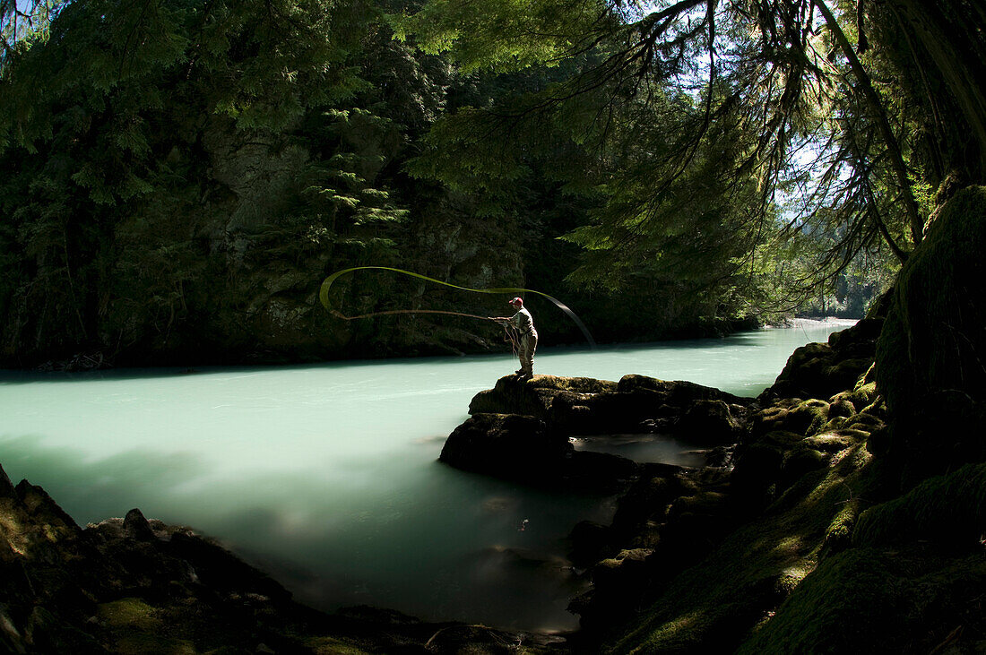 A man standing on a rock, casts into a river wearing waders while fly fishing in Squamish, British Columbia Squamish, British Columbia, Canada