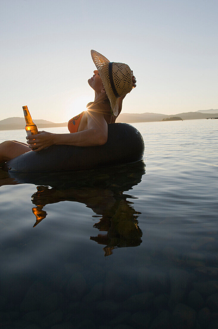 A young woman soaks in the last rays of the day while floating in a lake in an inner tube with a beer Sandpoint, Idaho, USA