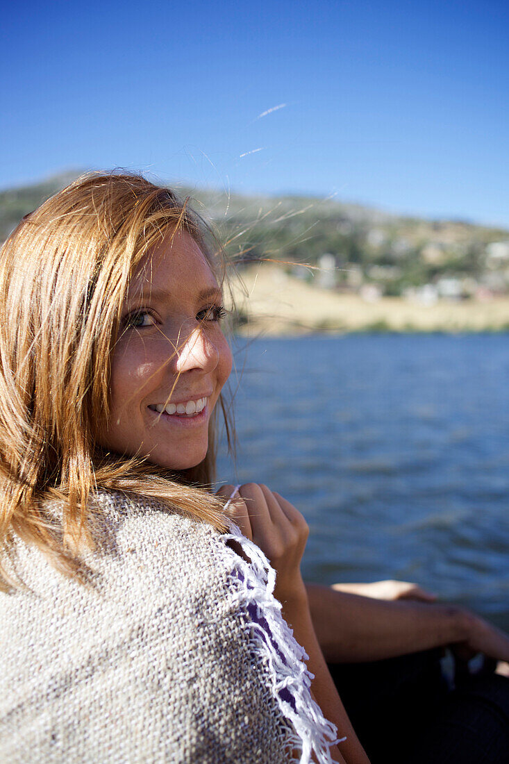 Young woman wrapped on a blanket sitting by a lake looks at the camera and smiles Julian, California, USA