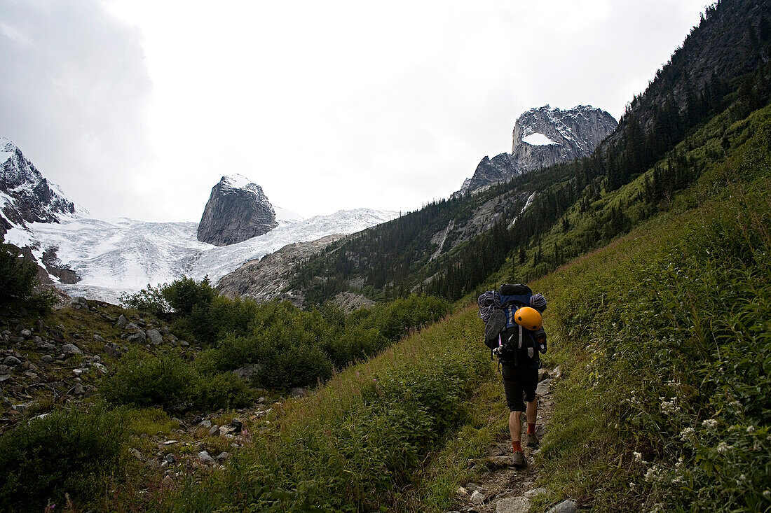 Man carrying a large backpack with a yellow climbing helmet attached to it hikes uphill towards a glacier British Columbia, Canada