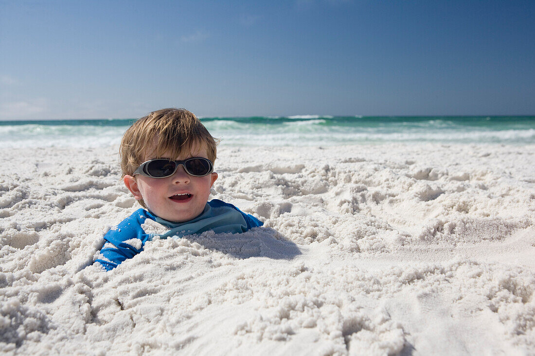 A little boy with sunglasses is buried in the sand up to his head with blue sky and ocean in the background Destin, Florida, USA