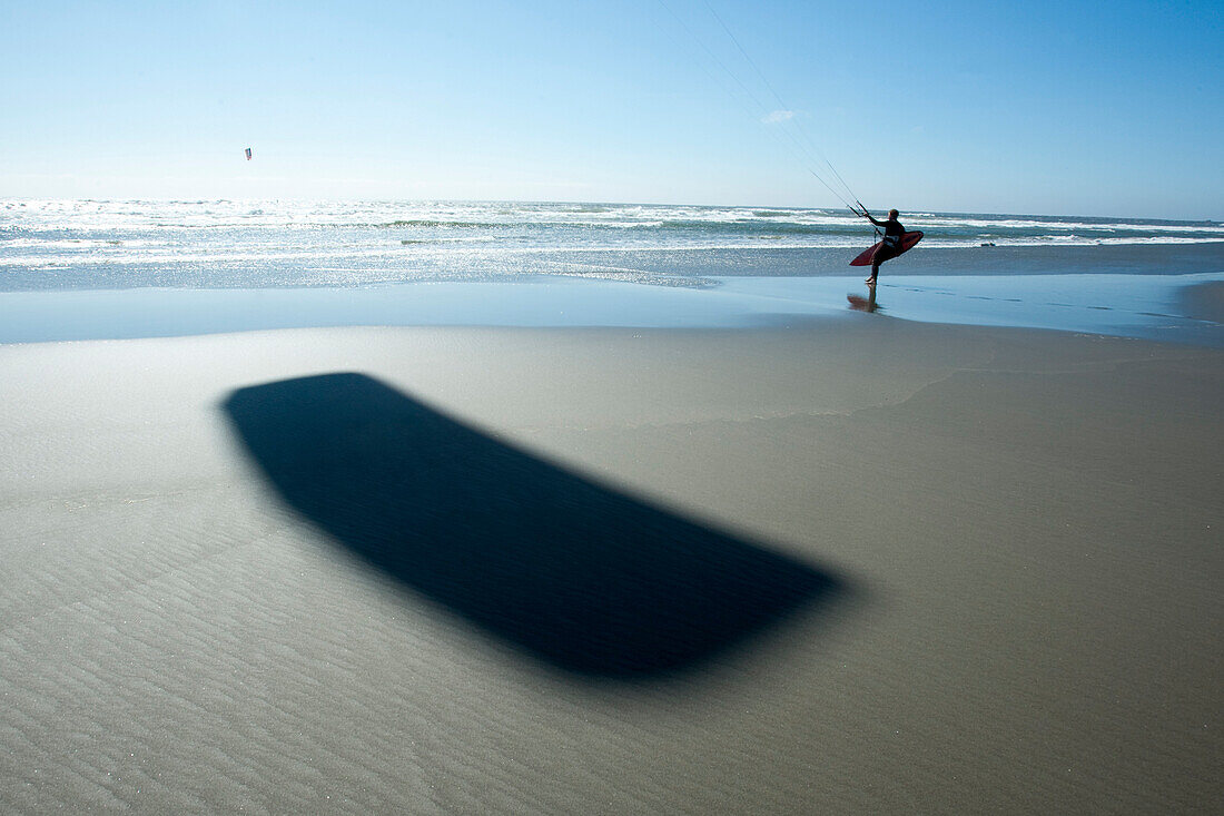 One male kitesurfer walking out into the ocean with the shadow of his kite prominent in the foreground Astoria, Oregon, USA