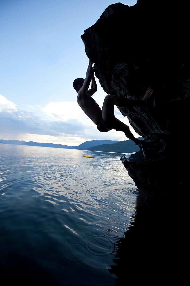 Young man free soloing up a rock with a kayak in the background in Idaho Sandpoint, Idaho, USA