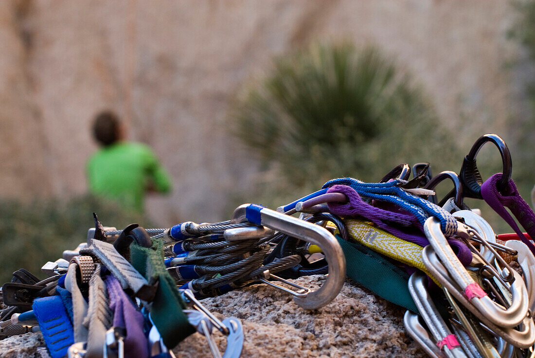 A rack of cams and nuts used for climbing rests a on a small boulder as a man belays a climber in the background Joshua Tree, California, USA