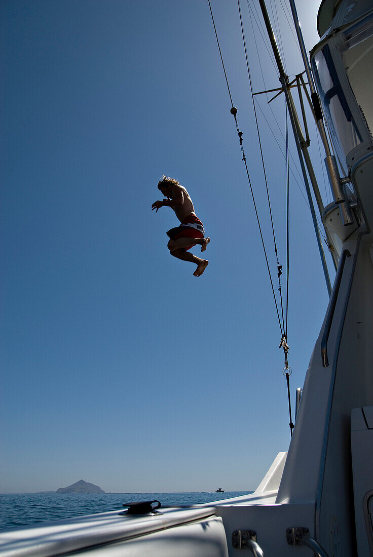 A young man jumps off of the top of a boat into the Pacific Ocean with an island in the background Channel Islands, California, USA