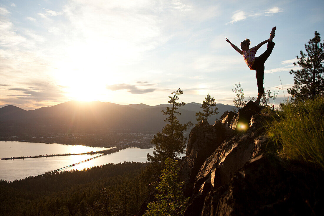 Young woman doing yoga at the top of a mountain overlooking a small town and large lake Sandpoint, Idaho, USA