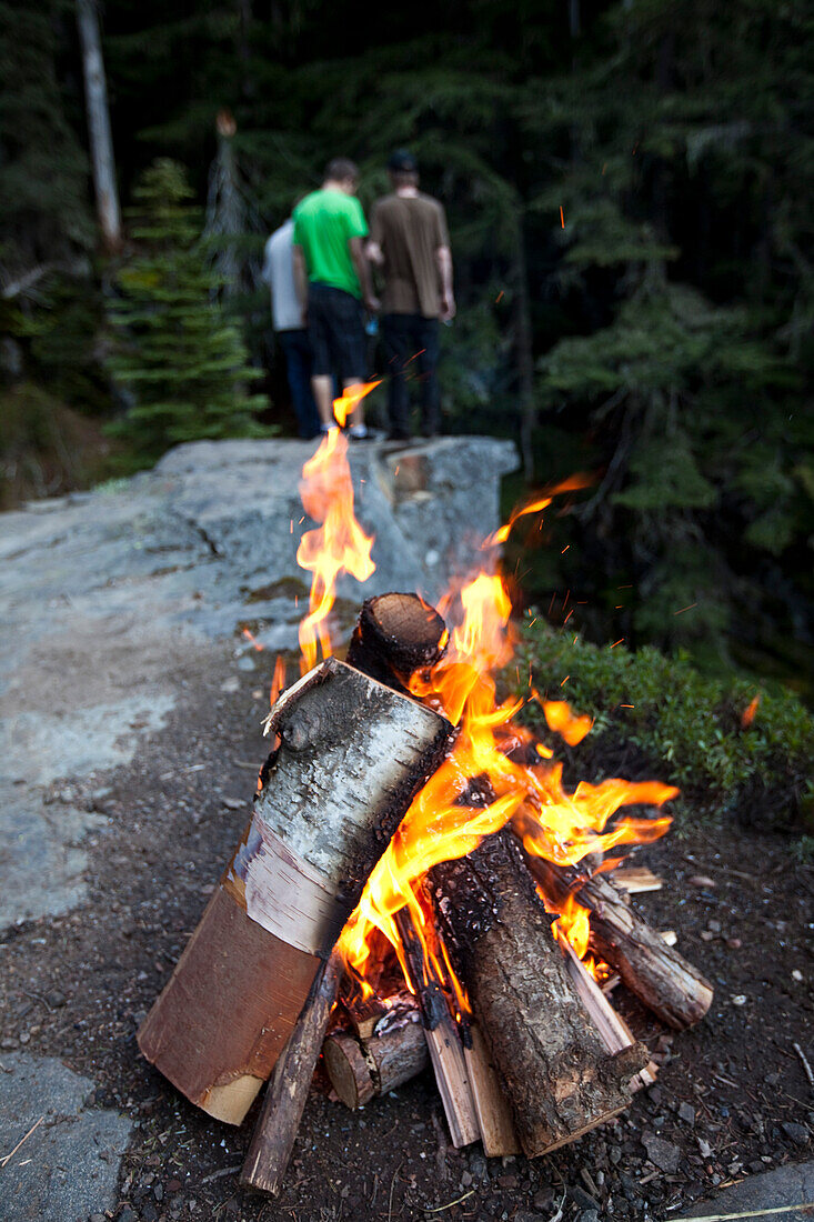 A campfire burns with three young men in the background looking over the edge of the cliff Sandpoint, Idaho, USA
