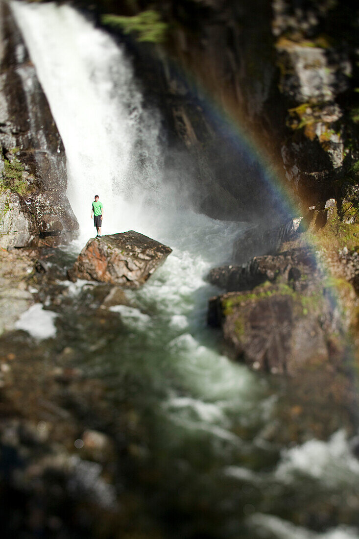 A young man stands at the base of a waterfall next to a rainbow with his arms open Sandpoint, Idaho, USA