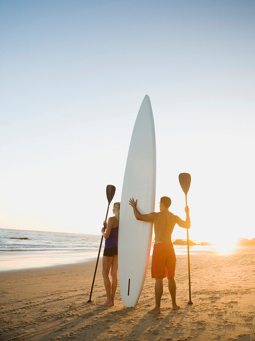 Couple standing on beach with surfboard and paddles, Newport Beach, CA, USA