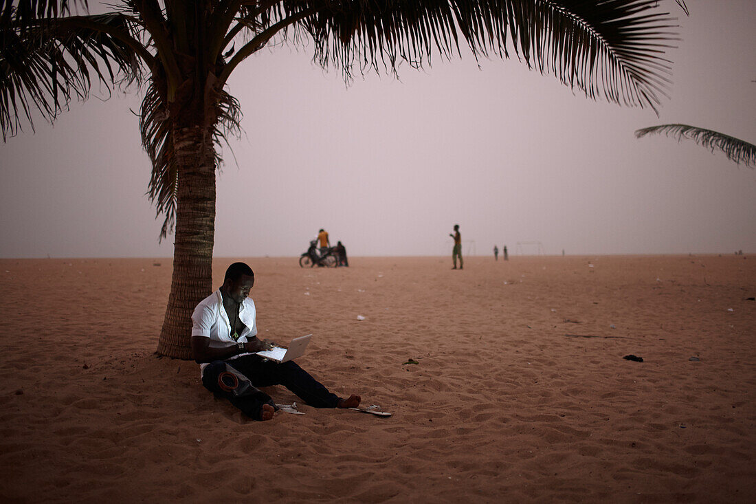 Mann arbeitet an einem Laptop unter einer Palme am Strand, Togbin Plage, Route des Peches, bei Cotonou, Benin