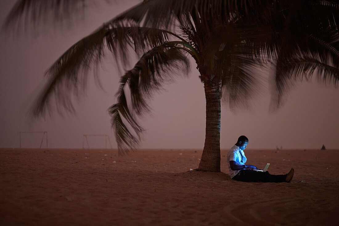 Mann arbeitet an einem Laptop unter einer Palme am Strand, Togbin Plage, Route des Peches, bei Cotonou, Benin