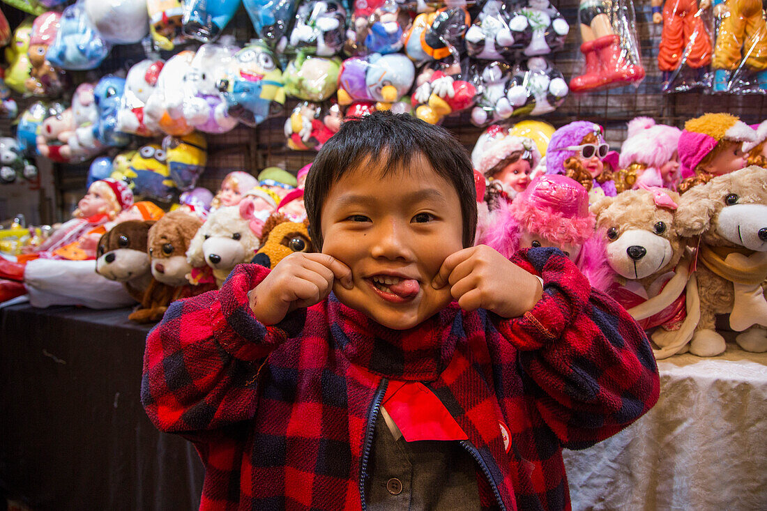 Kleiner Junge zieht Grimasse an einem Stand für Spielzeug am Temple Street Night Market Nachtmarkt, Tsim Sha Tsui, Kowloon, Hongkong, China