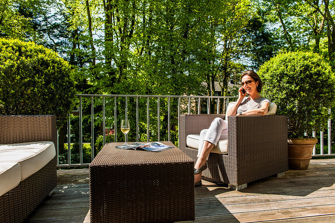 Frau mit Handy sitzt auf einem Balkon mit Loungemöbeln, Hamburg, Deutschland