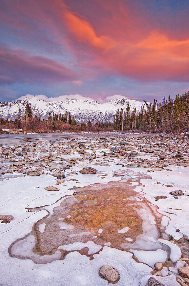 The Wheaton River In Early Winter With The Clouds Lit By Sunset Light. Snow Covered Mountains Seen In The Distance. Located Outside Of Whitehorse, Yukon. Rocks Surrounded By Ice In Foreground.