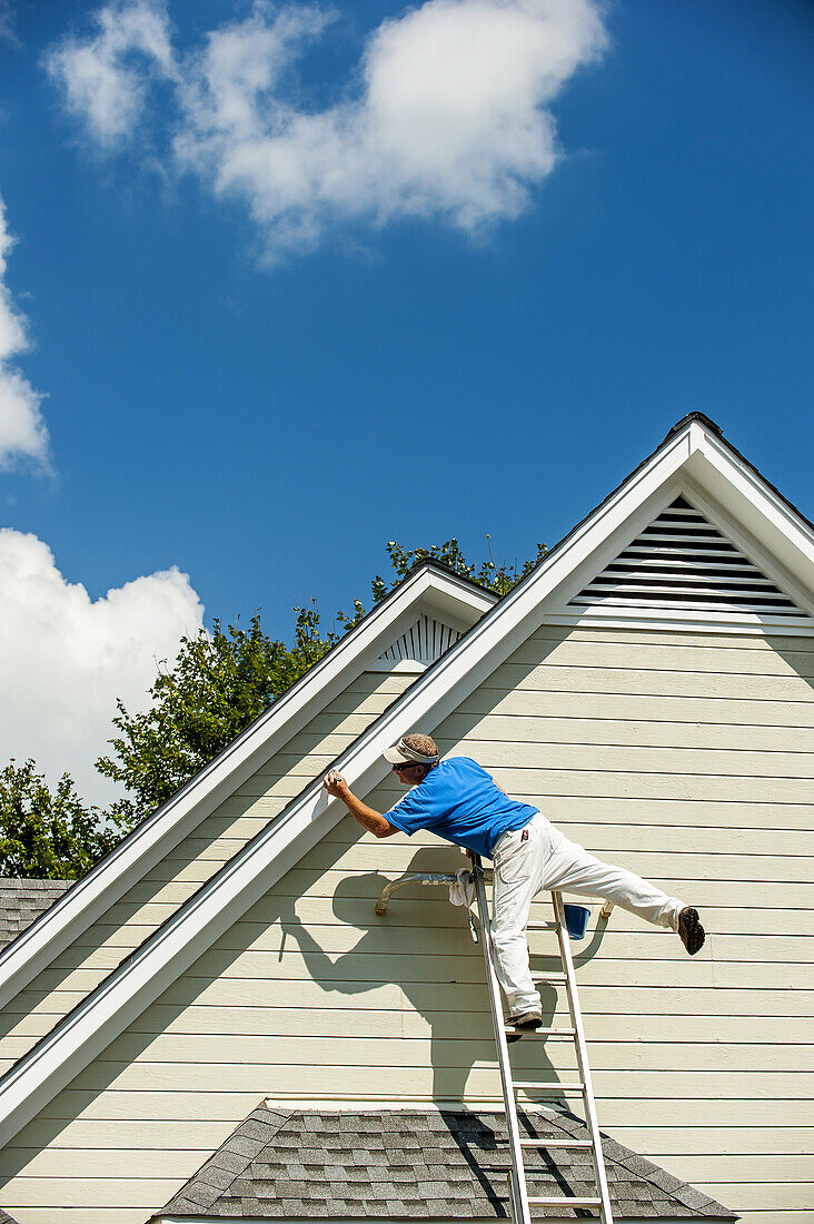 A 45 year old man on a ladder balancing on one leg clowning around while painting a house.