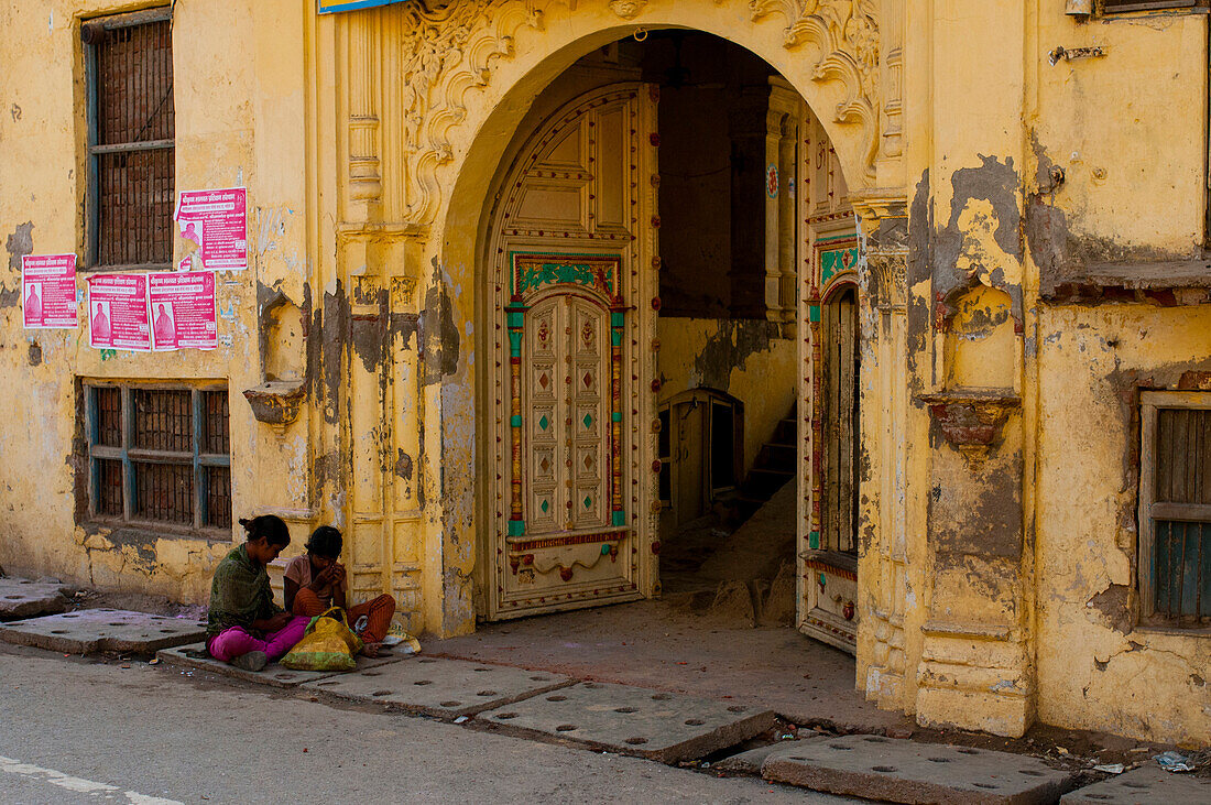 Two young women sits on the street's ground outside of a fancy entrance of a building in India