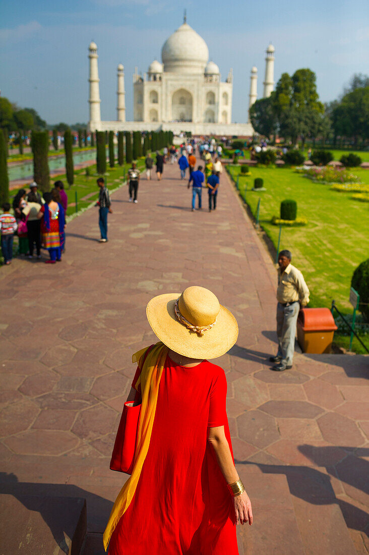 Elegant woman with red dress and large hat walks towards the Taj Mahal in the early morning light, Agra, India
