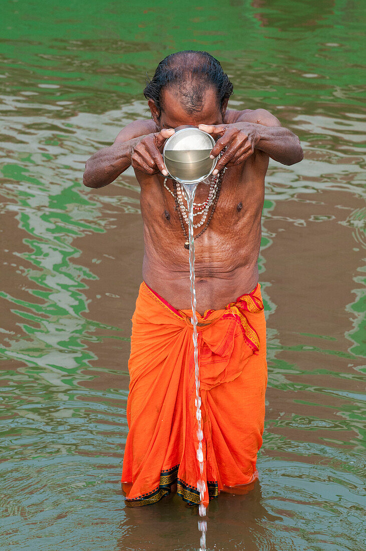 Man with an orange tunic holds a metal can throwing water in the holy Ganges river with a green background reflecting in the water, Varanasi, India